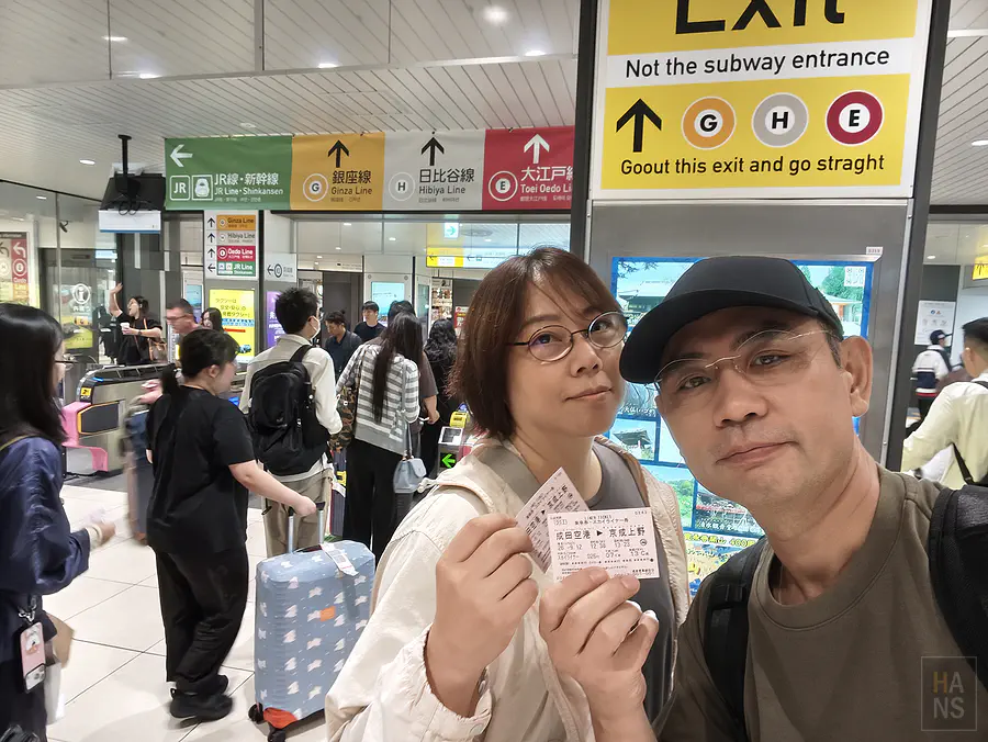 翰斯與Kay在京成上野站出口拿著Skyliner車票合照，轉乘JR與地鐵指標