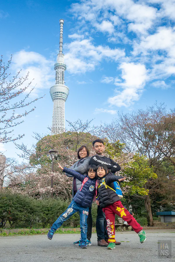 翰斯全家在晴空塔下公園草地擺出飛翔姿勢合照，背景為晴空塔與櫻花